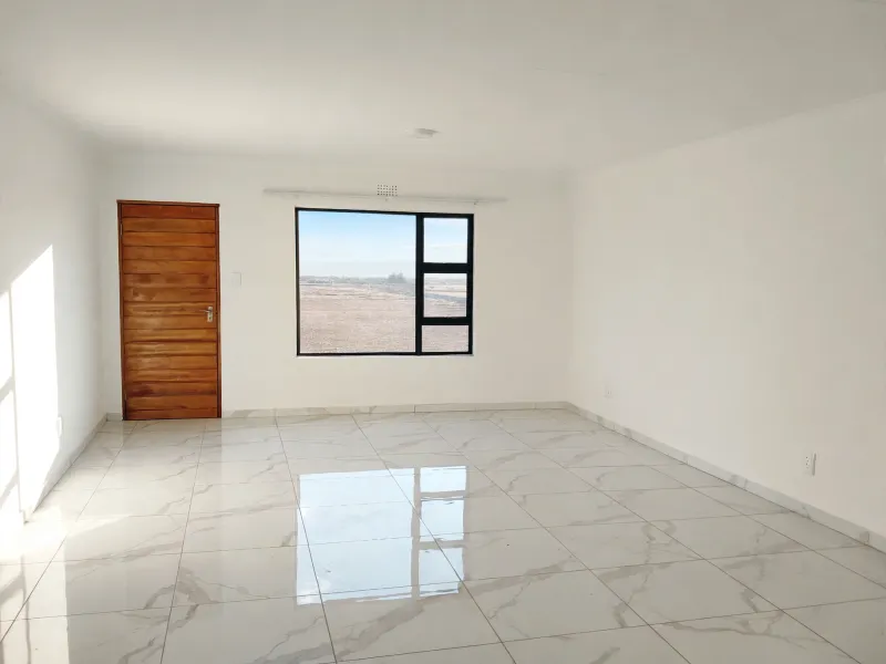 A bright, empty living area featuring polished marble-look floor tiles, a wooden door, and a large window with views of the surrounding landscape.