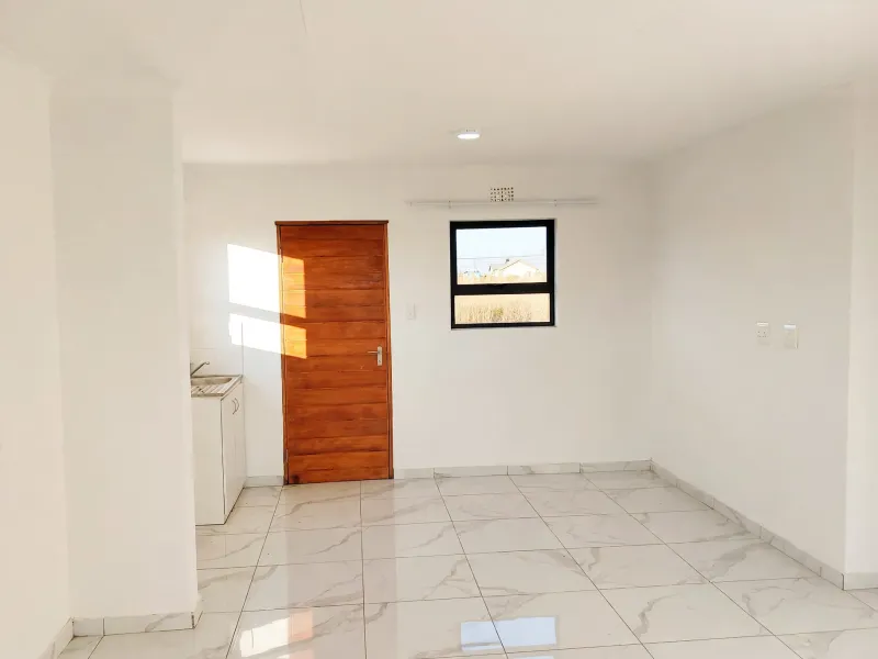 A bright, empty open-plan living area featuring modern marble-look floor tiles, a wooden door, and a compact kitchen sink unit.