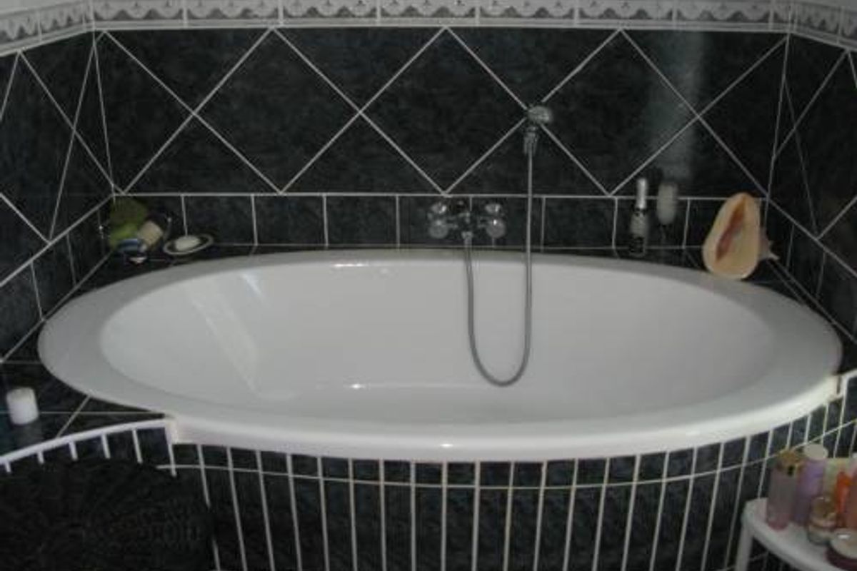 A bathroom featuring a built-in oval bathtub with a handheld shower mixer, surrounded by dark decorative wall and base tiling.