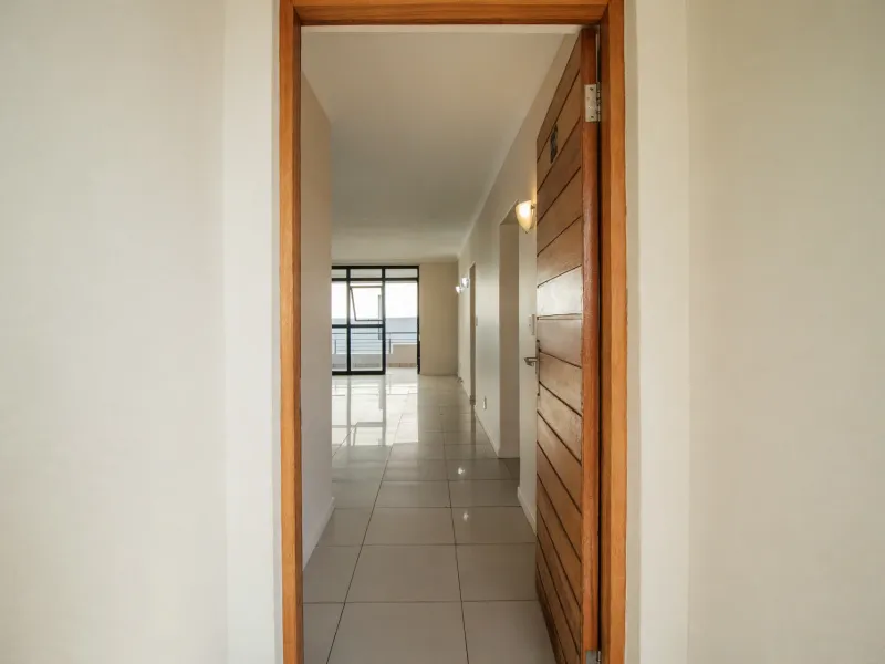 A bright, empty hallway with tiled flooring and wooden door, leading towards an open-plan living area with access to a balcony.