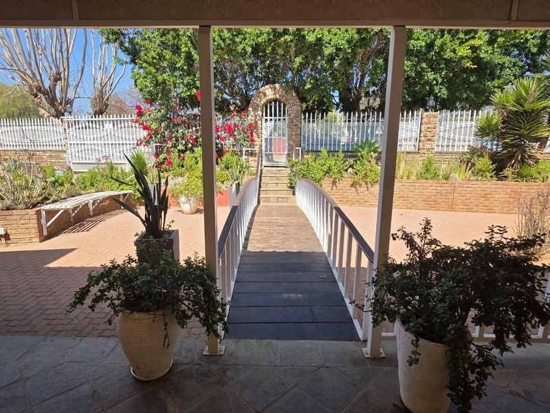 A covered patio area featuring brick paving and a raised wooden walkway leading to a stone archway and gated entrance in the garden.