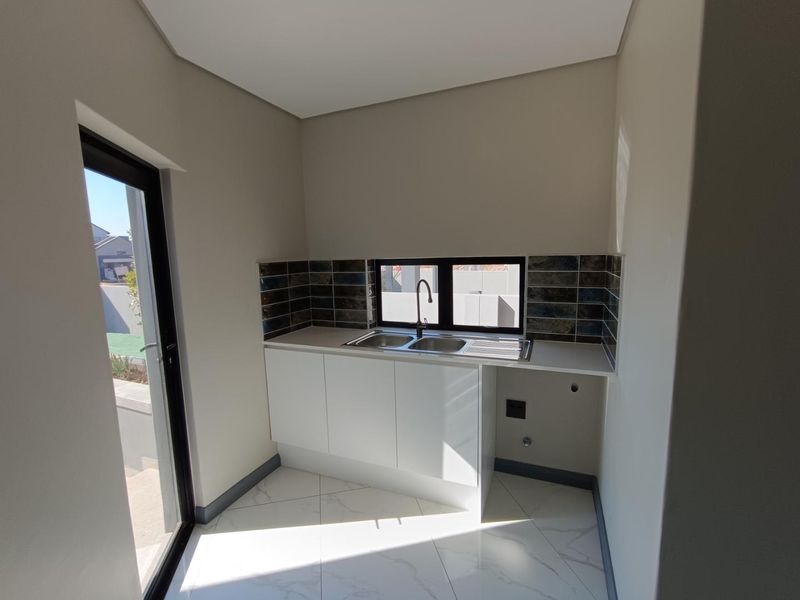 A clean, modern scullery featuring white built-in cabinetry, a double bowl sink, dark tiled splashback, and marble-look floor tiles with direct outdoor access.