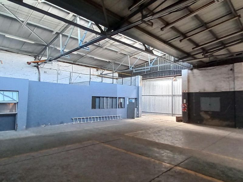 Spacious industrial warehouse unit featuring high exposed steel roof trusses, concrete flooring, and an internal office partition, suitable for various commercial or storage uses.