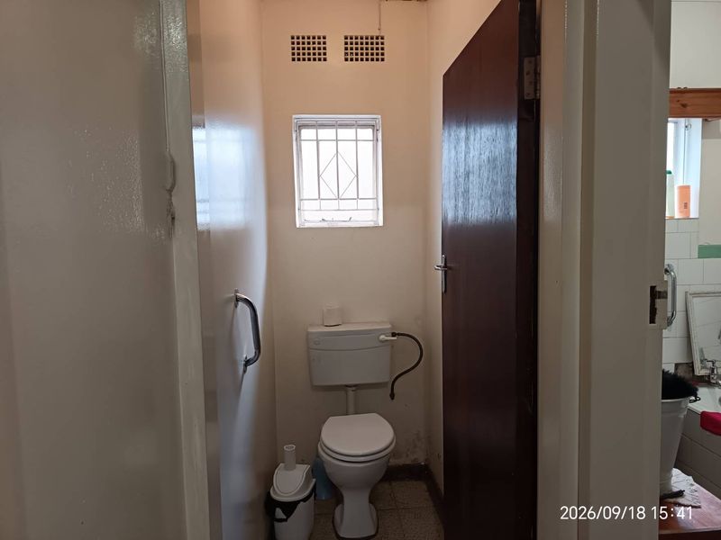 A compact, functional bathroom featuring a standard toilet, security-grilled window, and wall-mounted grab rail, requiring cosmetic updates to improve its overall condition.