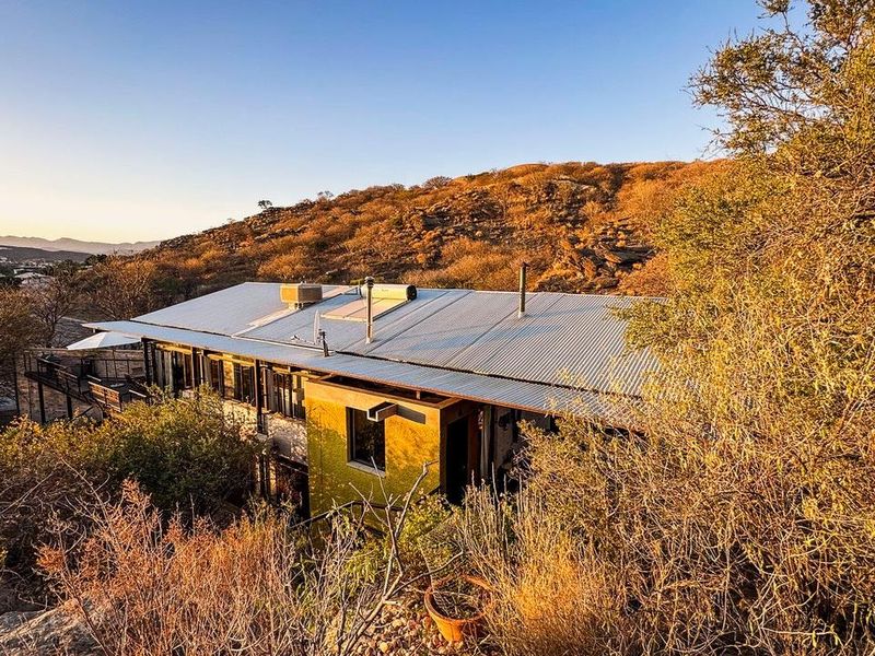 An elevated view of a hillside property featuring a corrugated metal roof, solar water heating system, and an attached outdoor deck area.