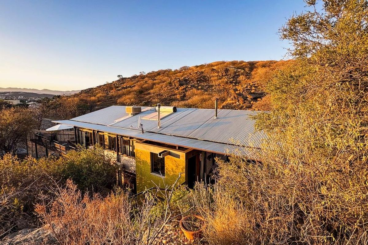 An elevated view of a hillside property featuring a corrugated metal roof, solar water heating system, and an attached outdoor deck area.