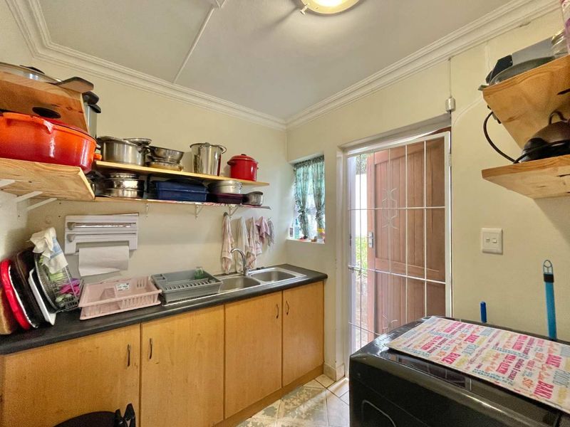 A functional scullery area featuring wooden cabinetry, a double sink, tiled flooring, and direct access to the exterior via a security gate.