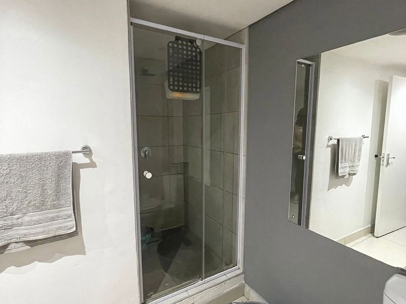 A compact bathroom featuring a glass-enclosed shower, wall-mounted mirror, and neutral tiling. The space requires minor maintenance to the shower base and finishes.