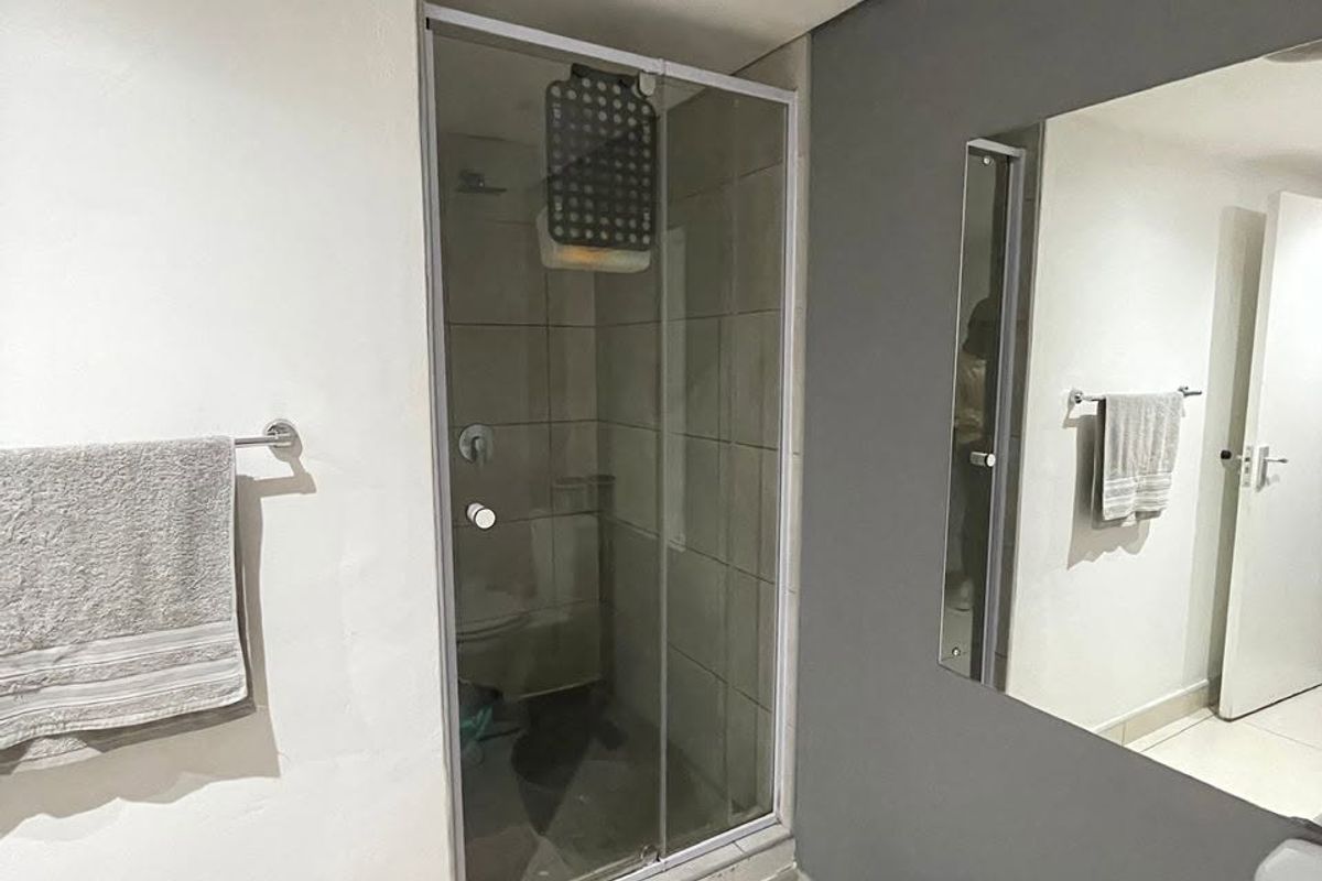 A compact bathroom featuring a glass-enclosed shower, wall-mounted mirror, and neutral tiling. The space requires minor maintenance to the shower base and finishes.