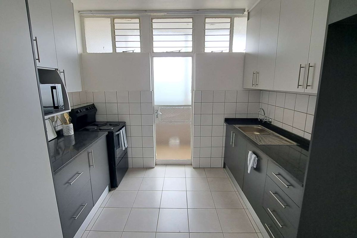 A functional galley-style kitchen featuring dark cabinetry, stone-look countertops, tiled splashbacks, and a built-in stove, with direct access to an exterior door.