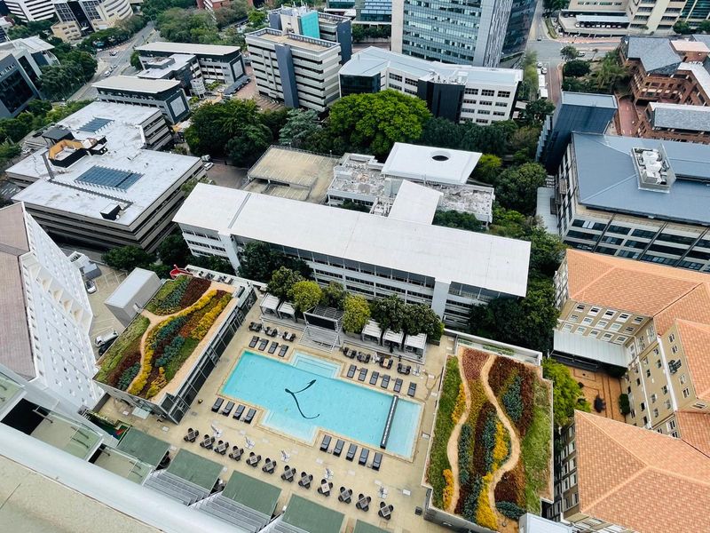 High-angle aerial view of a modern apartment complex featuring a swimming pool and landscaped rooftop gardens surrounded by urban infrastructure.