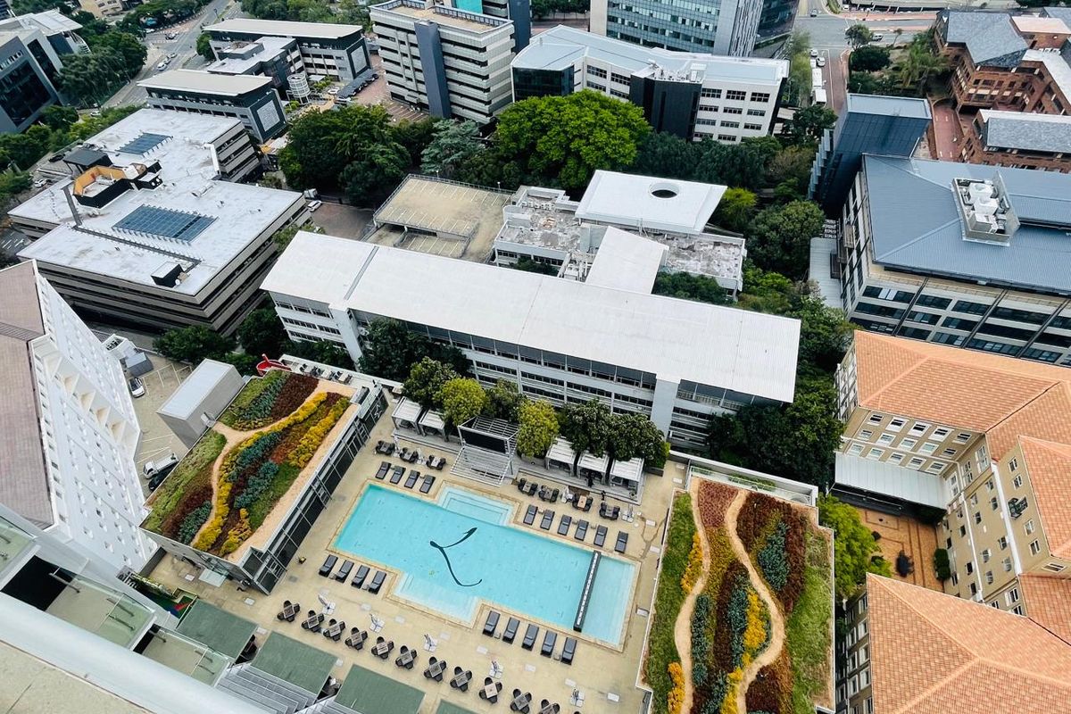High-angle aerial view of a modern apartment complex featuring a swimming pool and landscaped rooftop gardens surrounded by urban infrastructure.