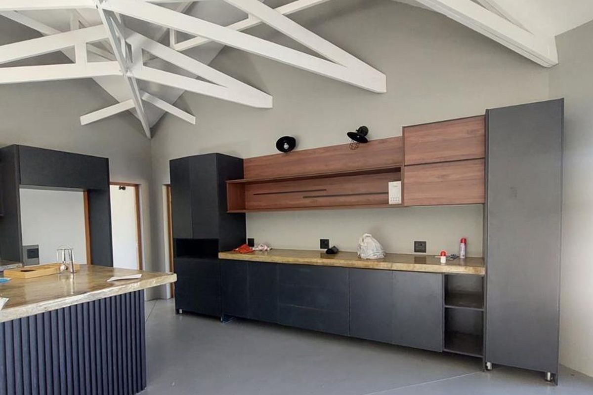 Modern kitchen featuring exposed white roof trusses, dark cabinetry, a central island with stone countertop, and sleek epoxy flooring in an open-plan layout.