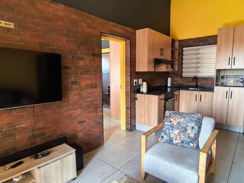 Open-plan living area featuring an exposed brick feature wall, tiled flooring, and a compact kitchen with built-in cabinetry and integrated appliances.
