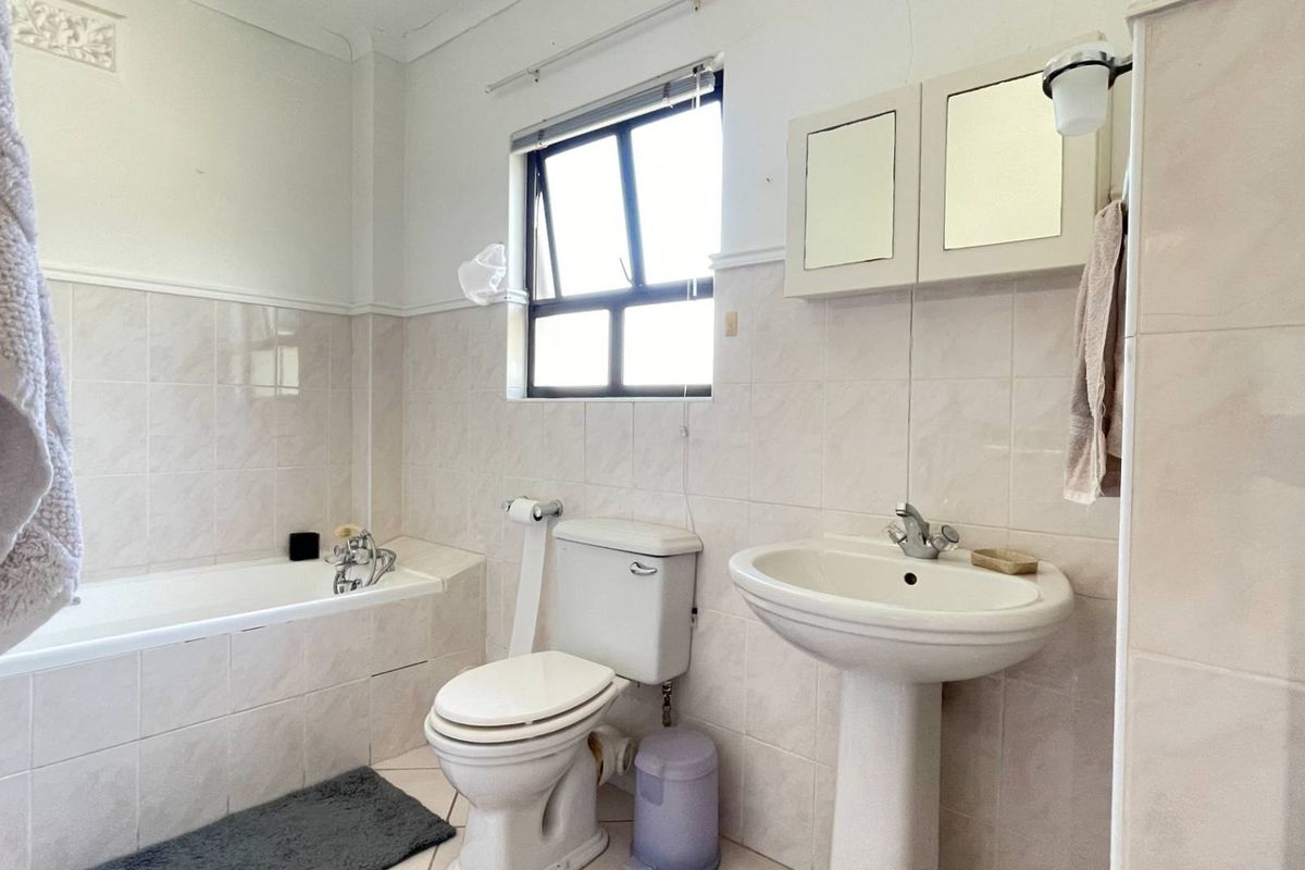 A functional bathroom featuring a built-in bathtub, pedestal washbasin, toilet, and wall-mounted medicine cabinet with neutral wall and floor tiling.
