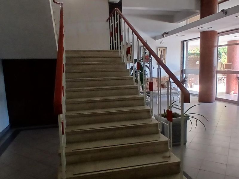 A spacious apartment building entrance lobby featuring a central staircase, tiled flooring, and large glass doors providing natural light.