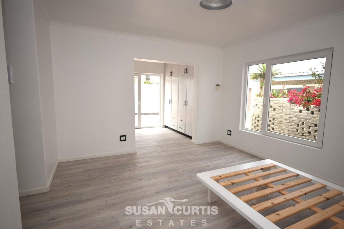 A bright bedroom featuring light wood-look laminate flooring, built-in cabinetry, and large windows overlooking a private garden area.