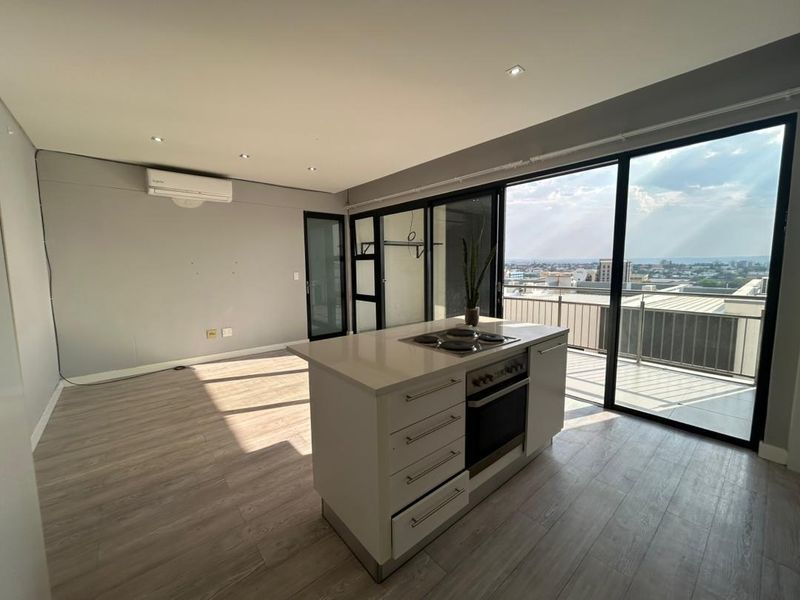 Modern open-plan living area featuring a central kitchen island with built-in appliances, laminate flooring, and large sliding doors opening to a balcony with city views.