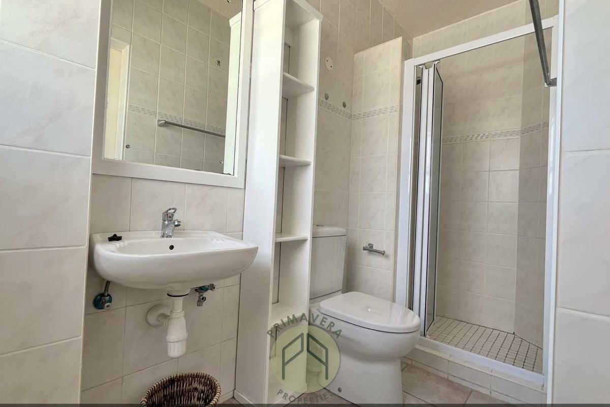 A functional bathroom featuring a wall-mounted basin, a built-in shelving unit, a standard toilet, and a glass-enclosed shower stall with tiled walls.