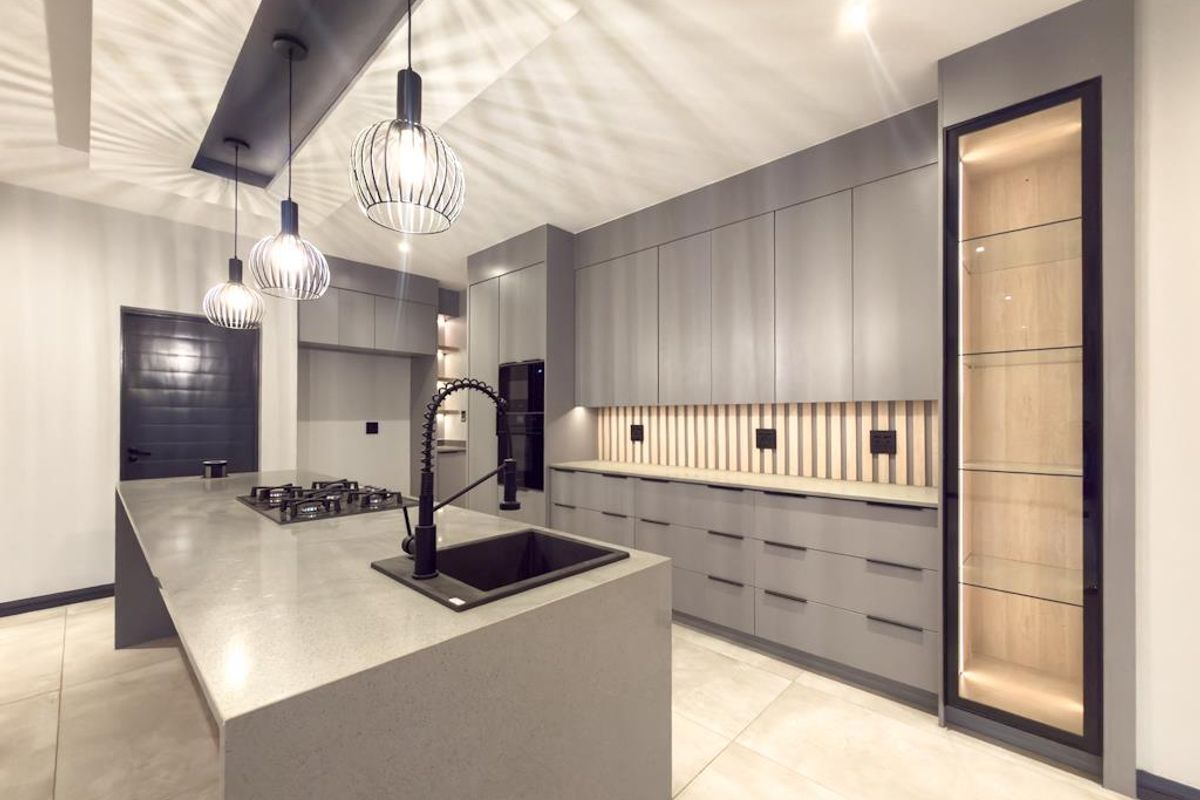 Modern kitchen featuring a large central island with a built-in gas hob, sleek grey cabinetry, integrated lighting, and a tall glass-fronted display cabinet.