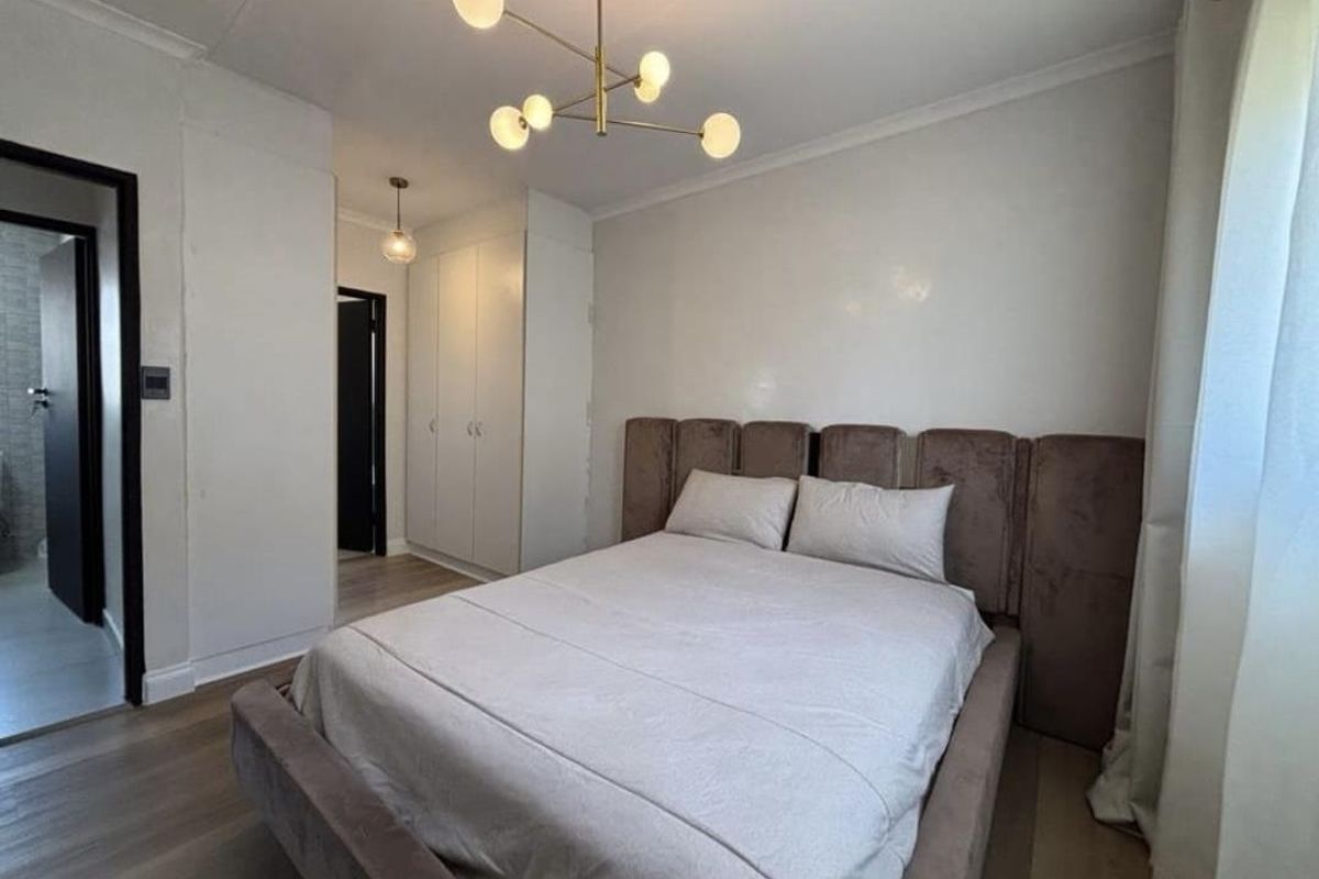 A well-maintained bedroom featuring built-in cupboards, modern light fixtures, and wood-look flooring, with an adjacent bathroom visible through the doorway.