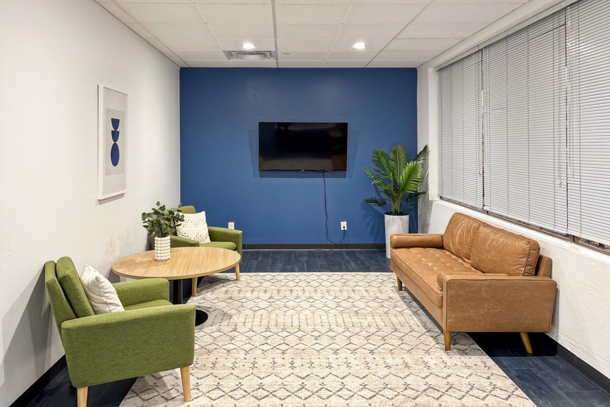 A modern office lounge area featuring a bold blue accent wall, suspended ceiling with recessed lighting, and large windows with venetian blinds.