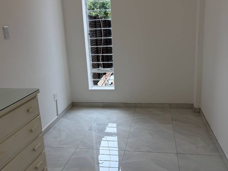 A bright, empty bedroom featuring modern tiled flooring and a tall window with security bars, providing a clean and neutral space.