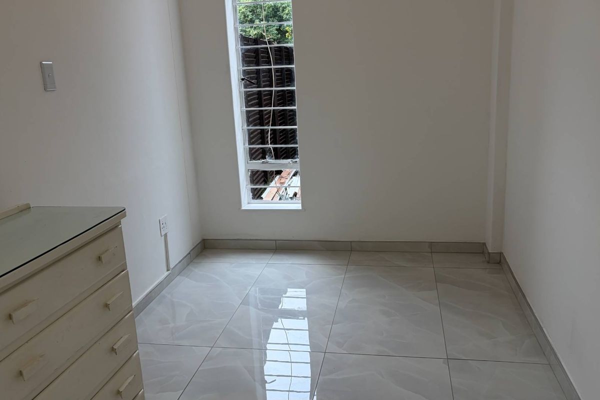 A bright, empty bedroom featuring modern tiled flooring and a tall window with security bars, providing a clean and neutral space.