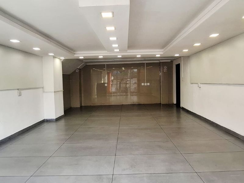 A spacious, empty commercial office area featuring modern tiled flooring, recessed ceiling lighting, and a glass partition wall.