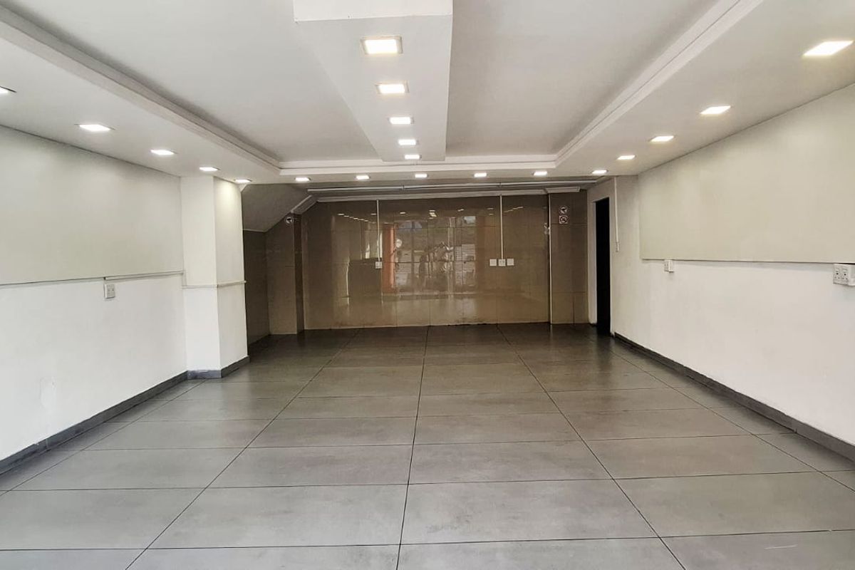 A spacious, empty commercial office area featuring modern tiled flooring, recessed ceiling lighting, and a glass partition wall.