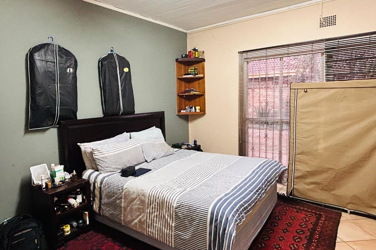 A bedroom featuring tiled flooring, a built-in corner shelf, and a large window with fitted blinds.