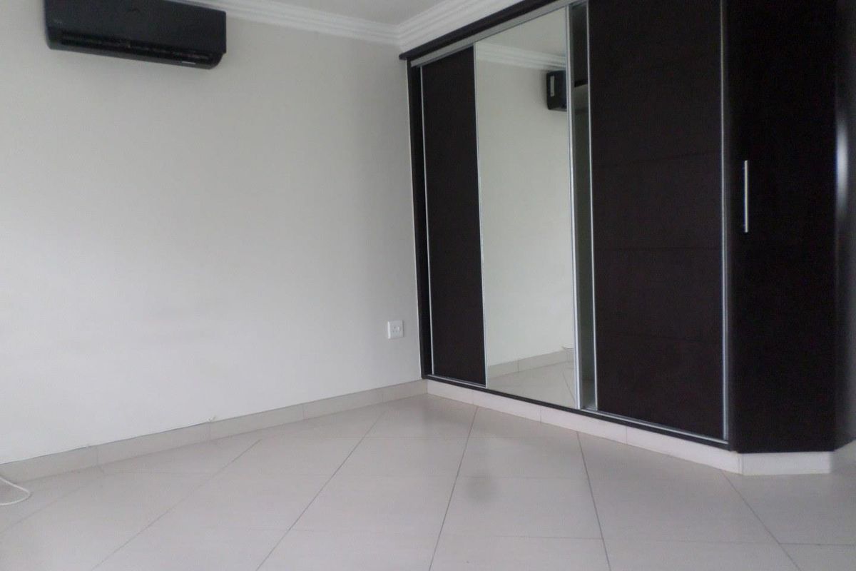 A clean, empty bedroom featuring tiled flooring, built-in sliding door wardrobes with a mirrored panel, and a wall-mounted air conditioning unit.