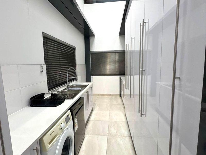 A modern, well-lit scullery featuring extensive white high-gloss cabinetry, stone countertops, a stainless steel sink, and integrated laundry appliance space.
