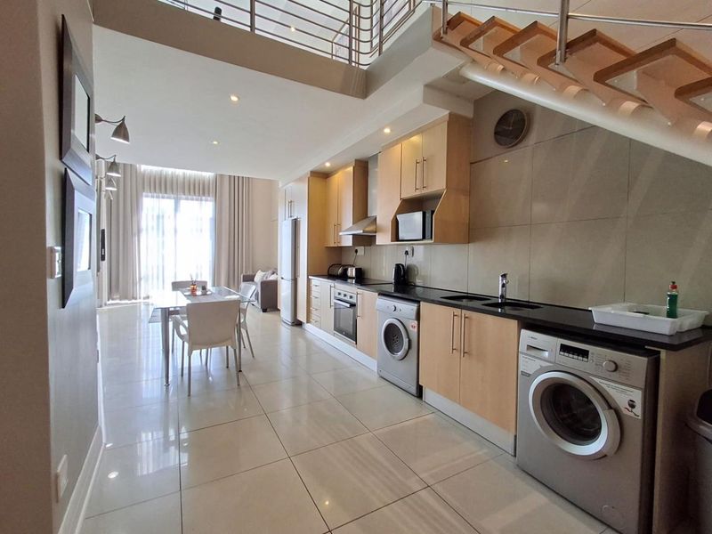 Modern open-plan kitchen and living area featuring light wood cabinetry, integrated appliances, sleek tiled flooring, and a staircase leading to an upper level.