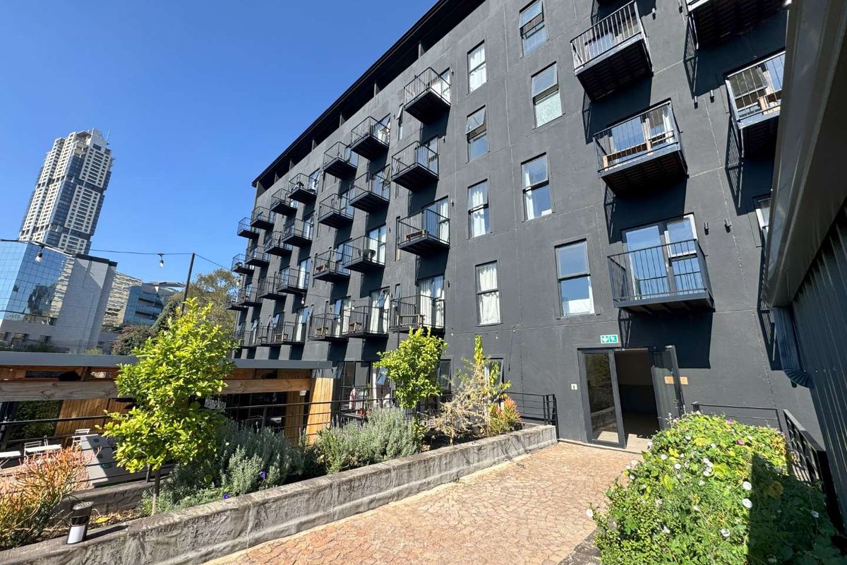 Exterior view of a modern apartment building featuring dark facade, multiple balconies, and a landscaped paved courtyard area.