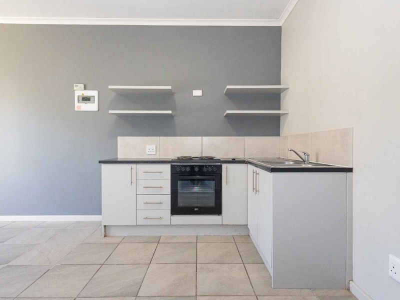 A compact, modern kitchen featuring built-in oven, electric hob, stainless steel sink, and floating shelves against a grey accent wall.
