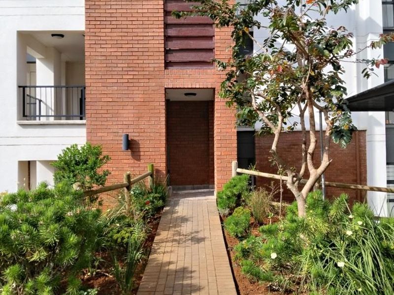 A well-maintained apartment building entrance featuring a brick facade, paved walkway, and landscaped garden area with timber fencing.