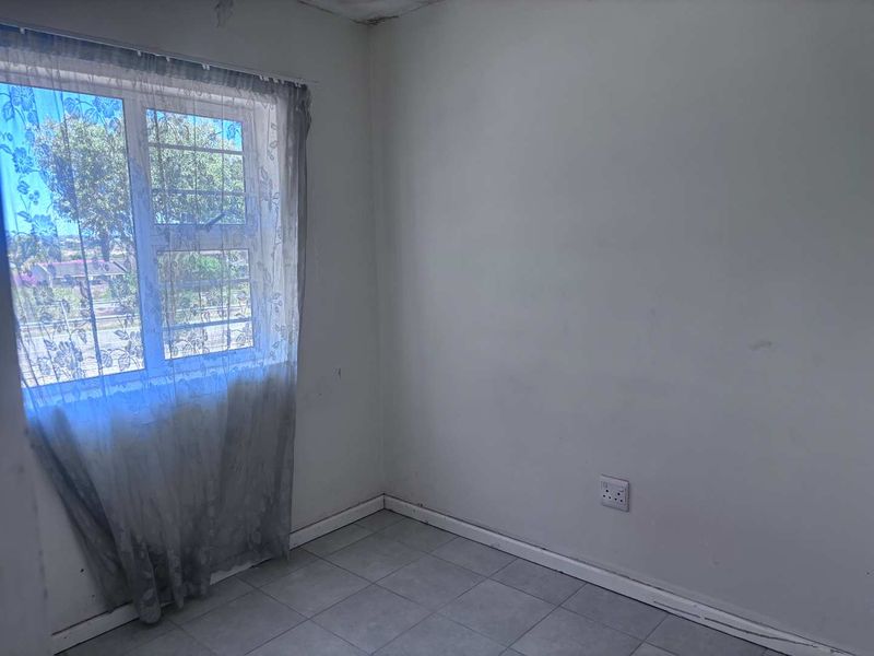 An empty bedroom featuring tiled flooring, an aluminium window with security bars, and visible signs of wear on the walls and ceiling.