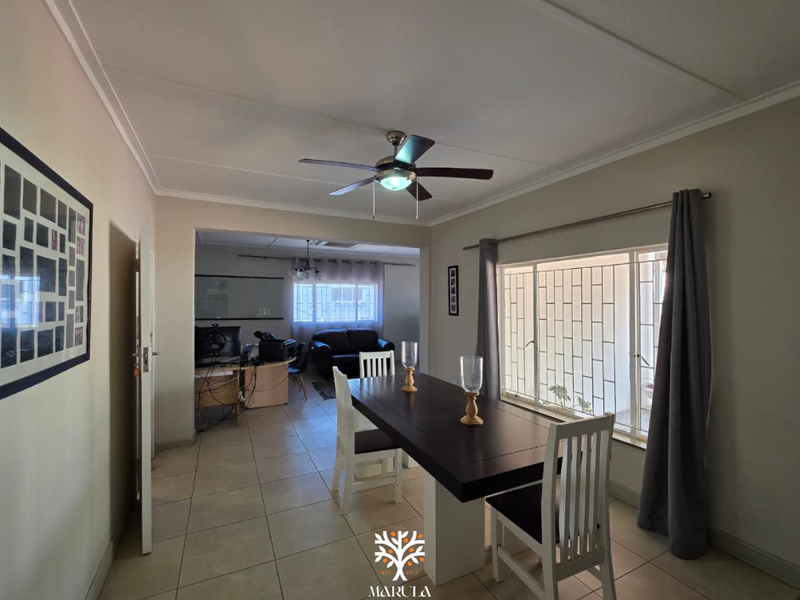 Dining room area featuring tiled flooring, a ceiling fan, and large windows with security bars, leading into an adjacent living space.