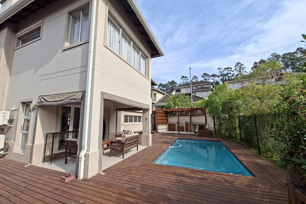 Rear exterior view featuring a swimming pool, timber decking, and a covered patio area with a built-in braai and pergola.