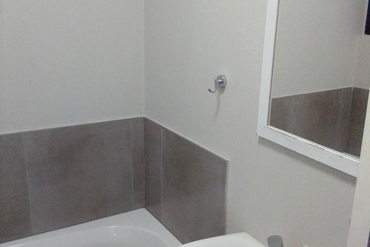 A clean, functional bathroom featuring a white pedestal basin, chrome mixer tap, bathtub with tiled splashback, and a wall-mounted mirror.