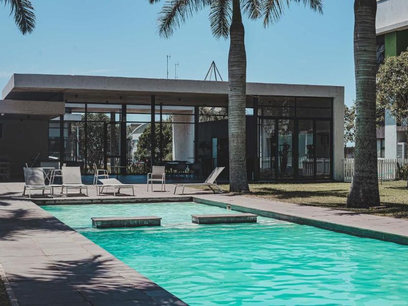 A well-maintained outdoor swimming pool area featuring a paved deck, mature palm trees, and a modern glass-walled clubhouse structure.