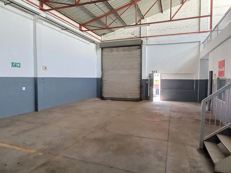 Spacious industrial warehouse unit featuring high ceilings, a large roller shutter door, concrete flooring, and an integrated staircase leading to an upper level.