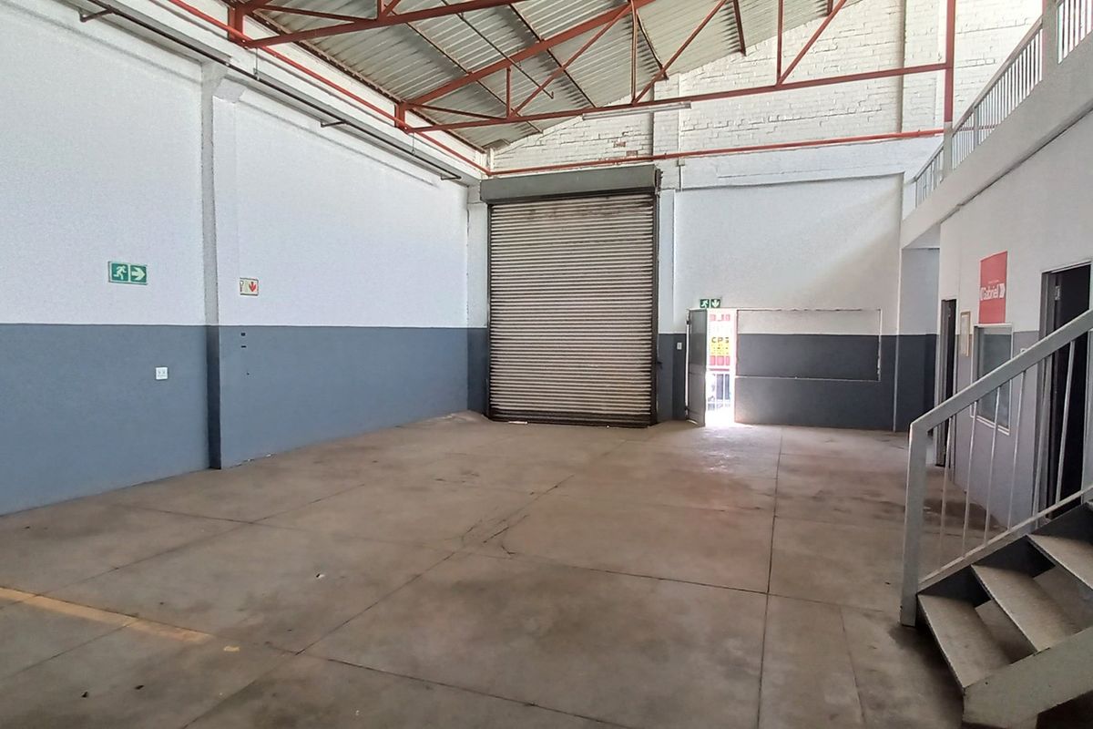 Spacious industrial warehouse unit featuring high ceilings, a large roller shutter door, concrete flooring, and an integrated staircase leading to an upper level.