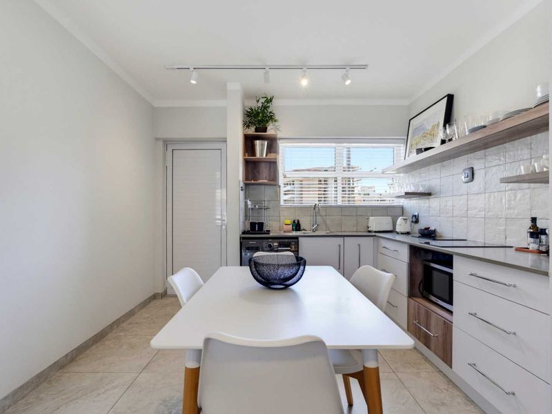 Modern open-plan kitchen featuring sleek white cabinetry, stone countertops, tiled backsplash, and integrated appliances, seamlessly flowing into the dining area.