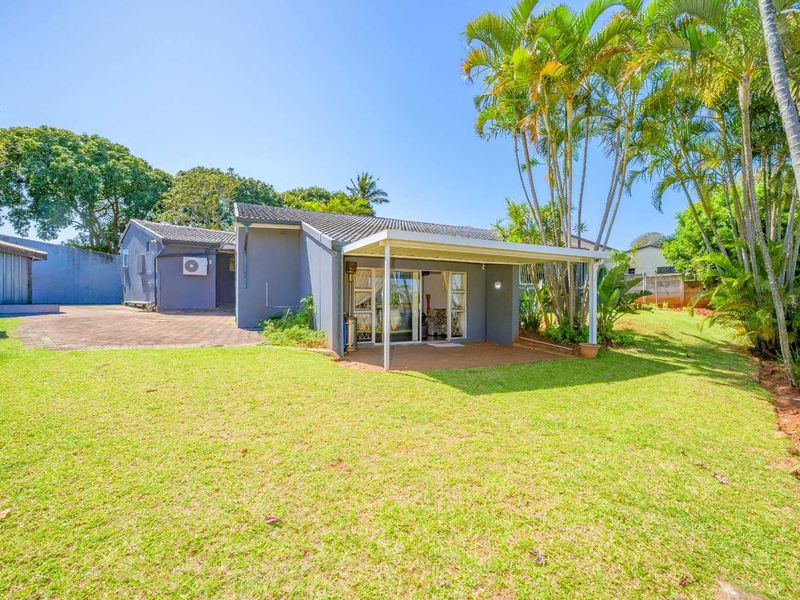 Rear view of the house featuring a covered patio, paved driveway, and a spacious garden with mature palm trees.
