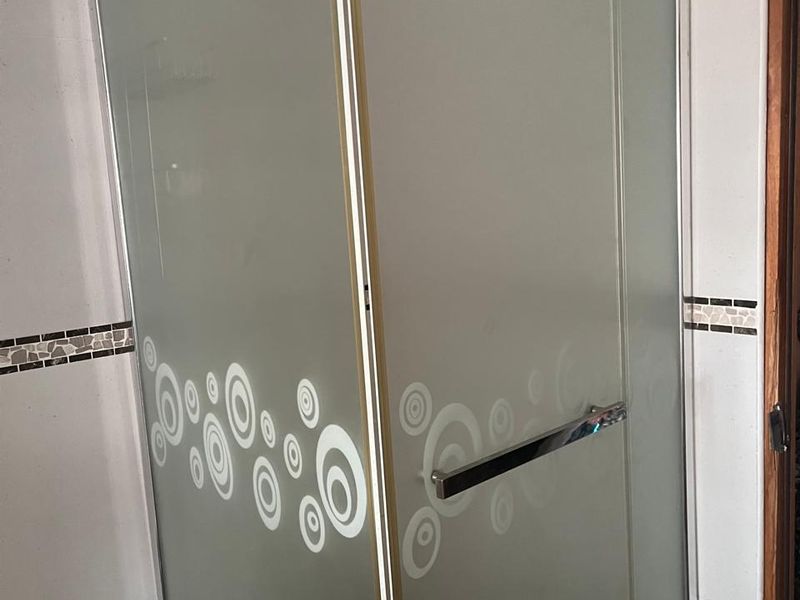A bathroom shower enclosure featuring frosted glass panels with a decorative circular pattern, chrome hardware, and tiled walls with a mosaic accent border.