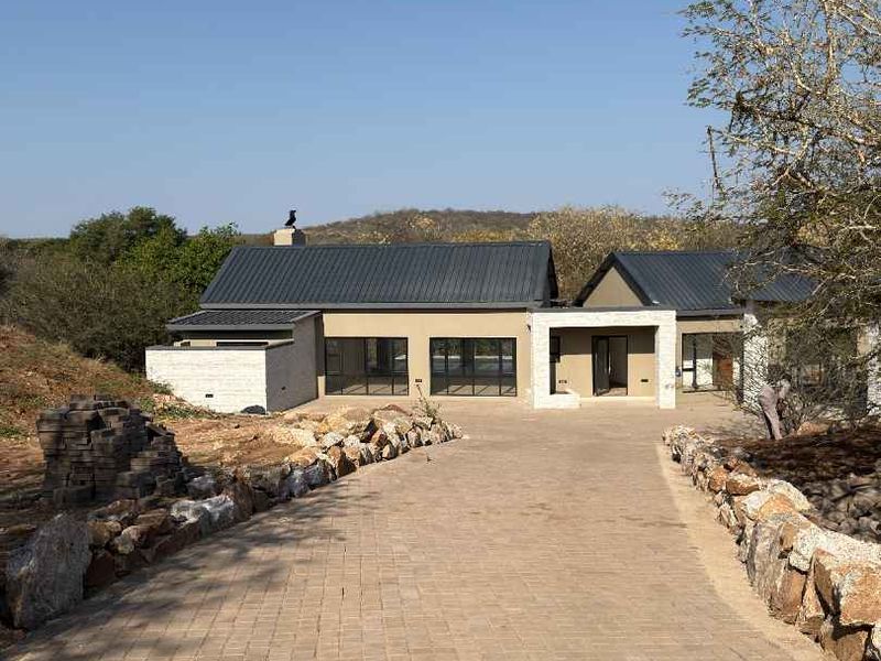 A modern single-story residence featuring a paved driveway, stone retaining walls, and a metal roof, set within a natural landscape.