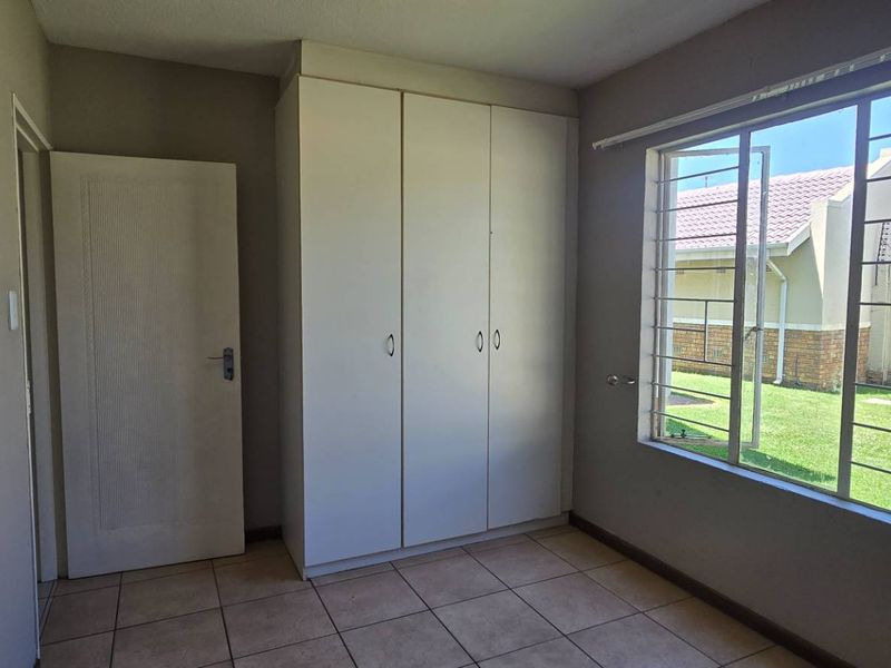 A bright, empty bedroom featuring tiled flooring, built-in cupboards, and a large window with security bars overlooking the garden.
