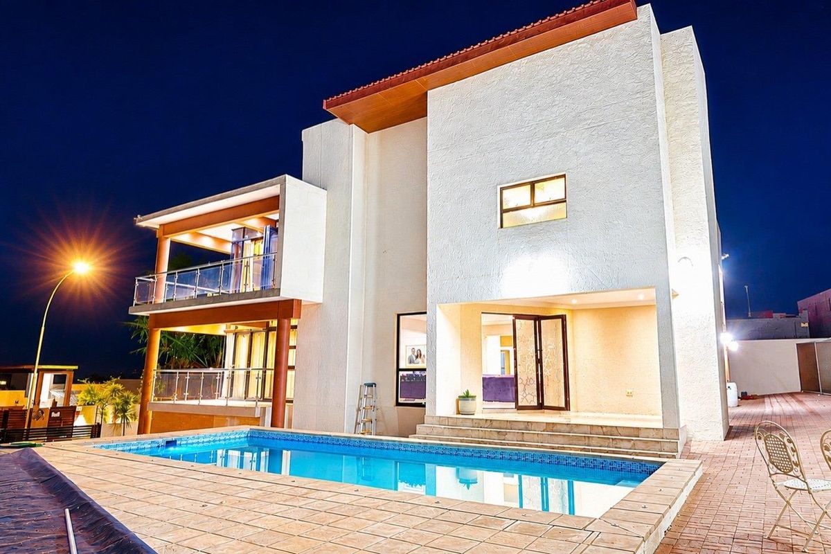 A modern two-story residence featuring a swimming pool, tiled pool deck, and upper-level balconies with glass balustrades, captured at twilight.
